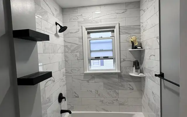 Bathroom remodel in Clifton, NJ with marble-look wall tile, tub and shower combination and black floating shelves