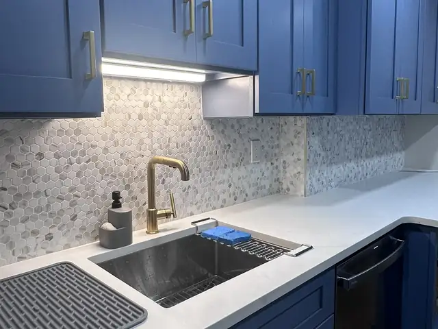 Blue kitchen cabinets with under-cabinet lighting, marble hexagon backsplash and brass faucet over a stainless sink